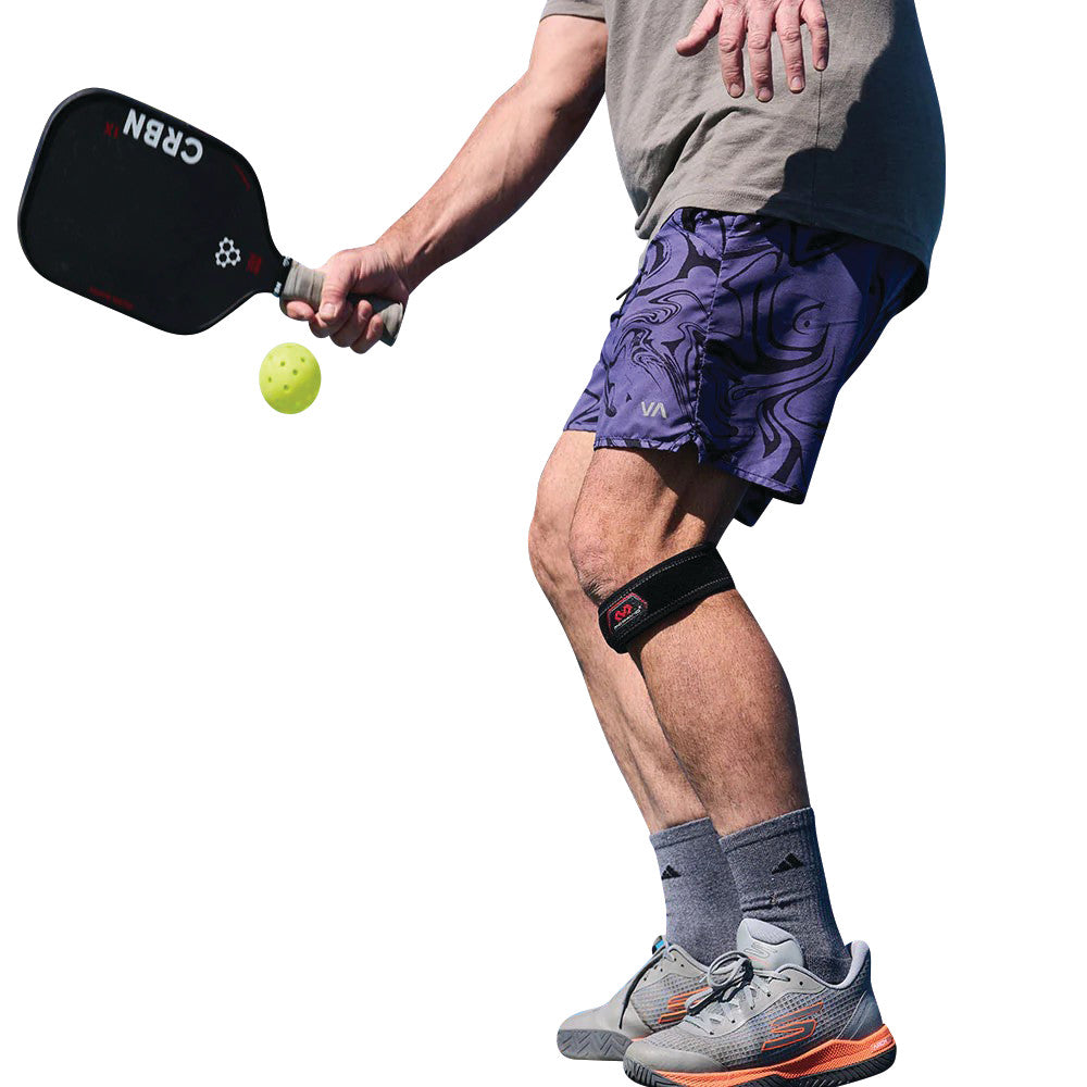 A person playing pickleball wears a McDavid Jumper's Knee Strap on their leg, holding a paddle and a yellow ball.