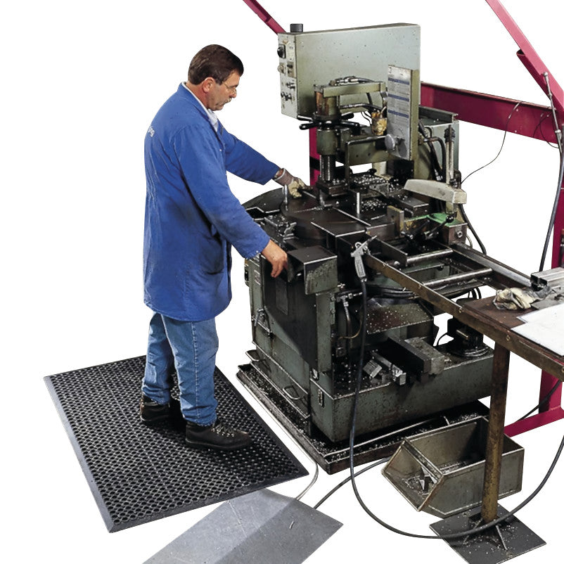 A man in a blue coat operates machinery while standing on a Sanitop Drainage Anti-Fatigue Mat, surrounded by industrial equipment.
