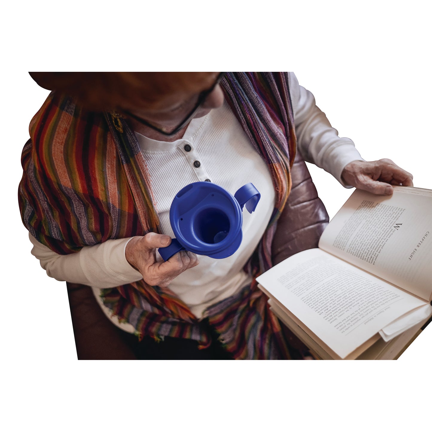 A person holds a blue Provale Cup while sitting and reading a book. They wear a white shirt and a colorful striped shawl.